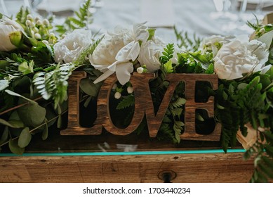 Love Written In Wood And Bridal Floral Arrangement With Eucalyptus And White Roses As Centerpiece On An Old Table