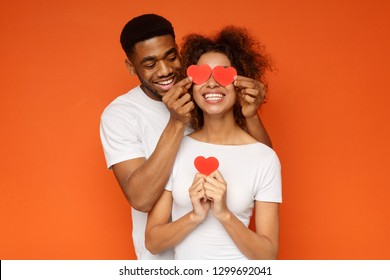 I Love You. Happy Young Man Is Covering Female Eyes With Surprise And Giving Her Heart Cards, Orange Studio Background