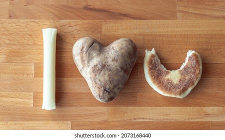 I Love You Message Spelled Out Using Food Items Including A Mozzarella String Cheese Stick, Heart Shaped Potato And An English Muffin Half With A Bite Out Of It On A Cutting Board Background.