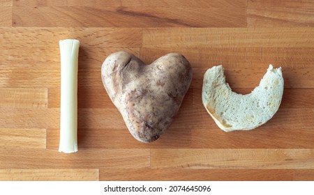 I Love You Message Spelled Out Using Food Items Including A Mozzarella String Cheese Stick, Heart Shaped Potato And An English Muffin Half With A Bite Out Of It On A Cutting Board Background.