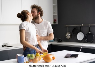 In Love Young Couple Kissing While Preparing Food At Kitchen. Husband Kiss His Wife While She Cut Oranges And Watching Videos Or Making Video Call Using Laptop. 