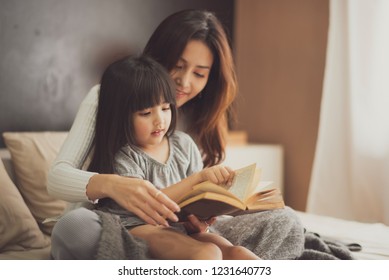 Love Of Young Mother And Daughter. Teaching Reading A Book On The Bed At Home. Mother And Daughter Look At The Book Feel Good And Happy