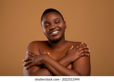 Love Yourself Concept. Happy Curvy African American Lady Embracing Herself, Pleased Black Millennial Female Standing With Closed Eyes And Smiling, Posing Naked Isolated Over Brown Studio Background