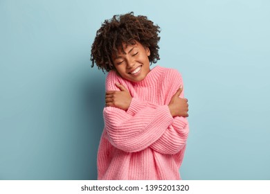 Love yourself concept. Photo of lovely smiling woman embraces herself, has high self esteem, closes eyes from enjoyment, likes her new comfortable soft pink sweater, tilts head, stands indoor - Shutterstock ID 1395201320