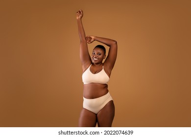 Love Yourself. Happy Chubby Black Woman Posing With Hands Raised Up, Showing Her Smooth Armpits, Cheerful African Female With Beautiful Skin Posing In Beige Lingerie At Brown Studio, Copy Space
