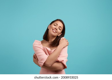 Love yourself. Happy smiling caucasian brunette wearing pink dress has high self esteem, keeps eyes closed with pleasure, feels enjoyment, embraces herself gently, isolated on blue background - Shutterstock ID 2008933025