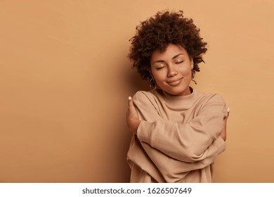 Love yourself, self acceptance. Gentle lovely Afro American woman crosses hands and embraces own body, tilts head and closes eyes, wears comforable brown sweater, models indoor. I love myself - Shutterstock ID 1626507649