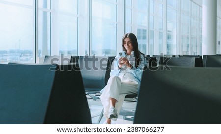 Lovely and adorable Caucasian lady sitting at airport waiting for departure. Charming and gorgeous young woman using smartphone online communicating. Traveling concept.