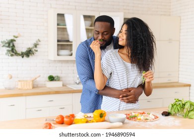 Lovely African American Couple Standing Next To Kitchen Counter, Hugging, Spending Leisure Time Weekend Together At Home, Girlfriend Looking At Boyfriend And Giving To Smell Herbs Mint, Cooking Pizza