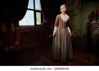 A Lovely Aristocratic Girl In An 18th Century Scottish Dress Stands In A Room With Classic Vintage Interior. The Heroine Of The Novel. 18th Century Style. Copy Space.