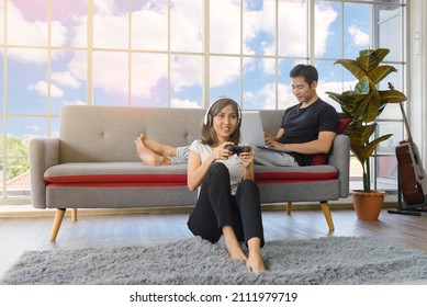 Lovely Asian Couple In Living Room At Home. Young Woman With Headphone Sit On Floor Playing Video Game, Man Lying On Couch Working With Laptop Computer In Background At Home. Love, Happiness, WFH