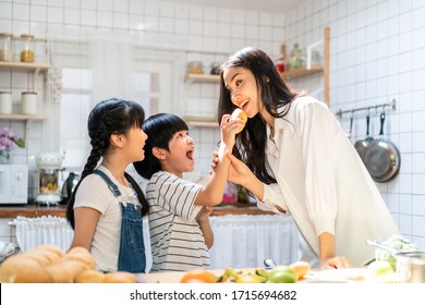 Lovely Asian Family Making Food And Son Give Mother Bread In Kitchen At Home. Smiling Mother And Children Standing At Cooking Counter That Food Ingredient Put On Table. Happy Family Activity Together.