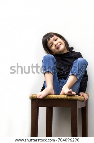 Lovely Asian little girl on black turtleneck and blue jeans sitting on bar stool.Girl in the good mood and poses while sitting on contemporary hi chair that made from wood and rattan.Gray background. 