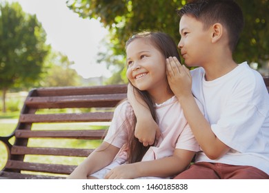 Lovely Asian Little Girl Smiling, Her Older Brother Whispering To Her Ear In The Park. Two Little Adorable Asian Children Enjoying Talking, Sitting On The Bench At The Park