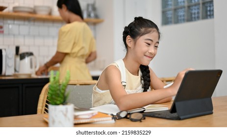 Lovely Asian Little School Girl Doing Homework, Using Tablet, Sitting At Dining Table While Her Mom Is Cooking Lunch In The Kitchen.