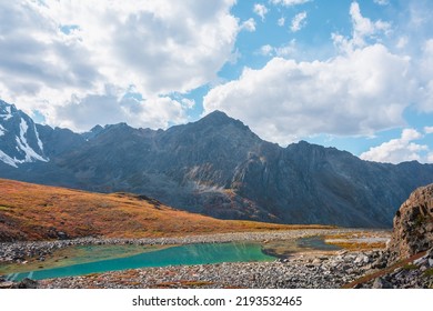 Lovely autumn landscape with turquoise mountain lake against high mountain top in sunlight under clouds in blue sky. Sunlit beautiful small alpine lake and large mountain range. Vivid autumn colors.