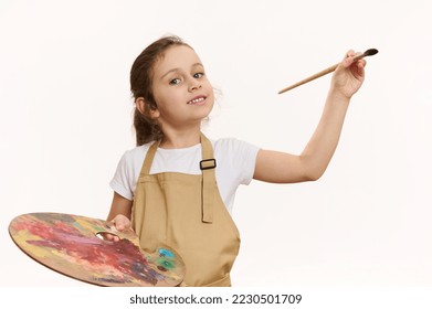 Lovely baby girl in beige apron, talented little painter with a paintbrush and palette of colorful paints, cutely smiling a toothy smile looking at camera, white background. Kids art development - Powered by Shutterstock