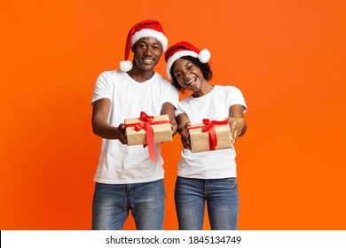 Lovely black couple showing their Christmas gift over orange studio background, copy space. Happy african american family wearing Santa hats and celebrating New Year 2021 together, holding present