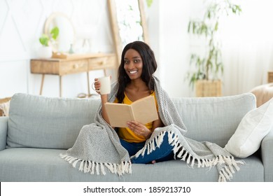 Lovely black woman reading book, covered with warm plaid, drinking hot aromatic coffee on sofa at home. Beautiful African American lady spending time indoors with captivating story - Shutterstock ID 1859213710