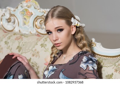 Lovely Blond Girl With Curly Hair Sitting On Vintage Sofa. Closeup Portrait Of Young Lady In Flowery Dress.