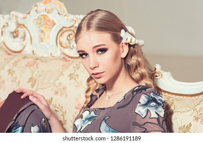 Lovely Blond Girl With Curly Hair Sitting On Vintage Sofa. Closeup Portrait Of Young Lady In Flowery Dress.