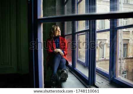 lovely blonde girl in a red jacket and blue jacket posing near the window with a purple frame in a house in St. Petersburg