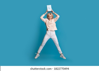 Lovely Blonde Lady Jumping On A Blue Studio Wall Holding Up A Box With Present While Smile At Camera