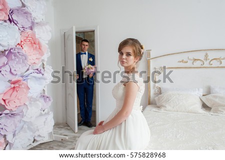 LOvely bride sitiing on the bed. Handsome groom in blue suit openening door with bouquet in his hands at the background