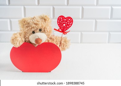 Lovely brown teddy bear toy sitting in wooden red heart shaped decor with carved heart decoration in its paw on white wooden table against tiled wall at home. Image with copy space, horizontal