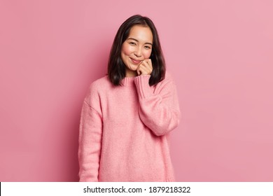 Lovely Brunette Woman With Eastern Appearance Keeps Hand On Face Has Gentle Smile Looks Satisfied At Camera Has Dimples On Rouge Cheeks Poses Against Pink Background. Cute Glad Asian Girl Indoor