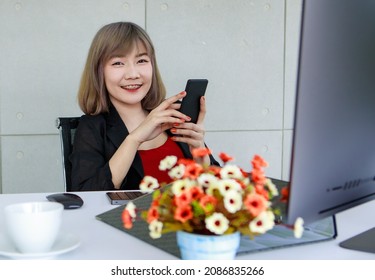 Lovely Business Asian Woman As Company Secretary And Admin Enjoy Online Amusement On Mobile Phone On Coffee Break Relaxation At Modern Office Working Desk Of Computer Monitor And Beautiful Flower.