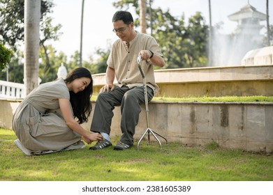 A lovely and caring young Asian daughter is helping her senior dad put shoes on, tie shoelaces while relaxing in a green park outdoors together. - Powered by Shutterstock