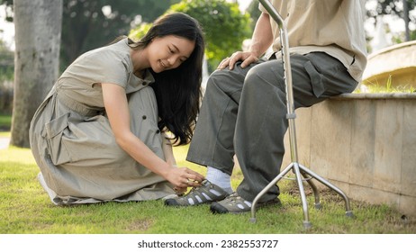 A lovely and caring young Asian daughter is helping her senior dad put shoes on, tie shoelaces while relaxing in a green park outdoors together. close-up side view image - Powered by Shutterstock