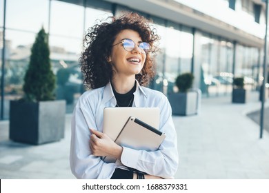 Lovely caucasian businessperson with curly hair posing outside with a tablet and laptop while wearing eyeglasses in a sunny day - Powered by Shutterstock