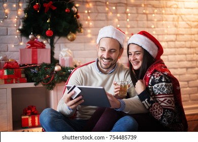 Lovely Charming Couple Sitting On A Floor With Santa Hats Hugged While Shopping Online On A Tablet And Drinking A Tea For Chrismas Holidays.