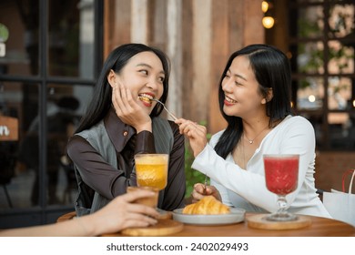 A lovely and cheerful Asian girl is feeding food to her female friend, enjoying eating food at a restaurant or cafe together. People and food concepts - Powered by Shutterstock