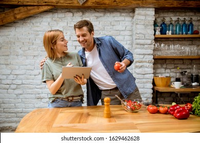 Lovely Cheerful Young Couple Cooking Dinner Together, Looking Recipe At Digital Tablet And Having Fun At The Rustic Kitchen