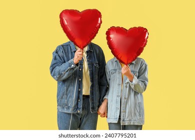 Lovely couple covering faces with heart-shaped balloons on yellow background. Valentine's Day celebration