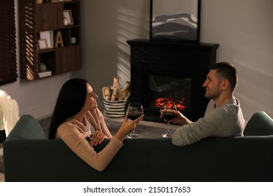 Lovely Couple With Glasses Of Wine Spending Time Together On Sofa Near Fireplace At Home