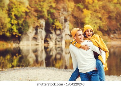 Lovely couple having fun together in nature. Fashion couple enjoying autumn. Boyfriend carrying his girlfriend on piggyback.Young couple walking near lake in autumn.Fashion, lifestyle and autmn mood.