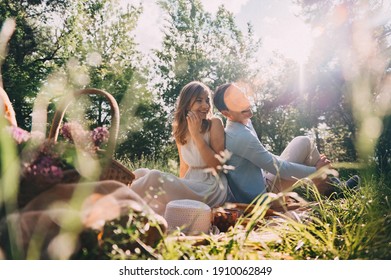 Lovely Couple In Love Organized A Picnic In The Park Wicker Basket With Flowers And Food On The Bedspread. Happy Lovers Laugh And Eat At The Picnic. Romantic Date