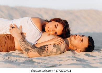 Lovely couple lying on the sand at sunset. A girl with wavy hair hugs her tattooed boyfriend