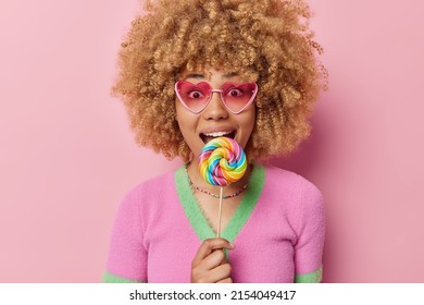 Lovely curly haired millennial girl bites colorful caramel candy on stick wears heart shaped sunglasses and casual jumper has sweet tooth isolated over pink background. Unhealthy food concept