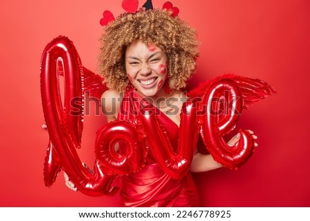 Lovely curly haired woman holds letter shaped balloons invites you on Valentines Day party wears dress and wings behind back expresses positive emotions isolated over red background. 14th February