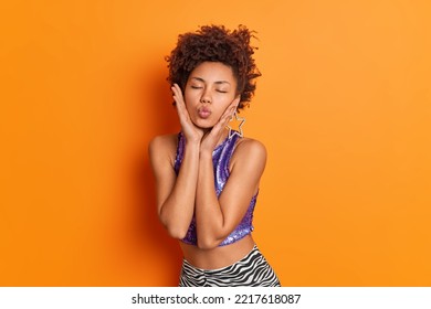 Lovely Curly Haired Woman Keeps Lips Folded Touches Cheeks Wears Purple Top And Leggings Expresses Love And Care Has Romantic Mood Isolated Over Vivid Orange Background. Human Facial Expressions