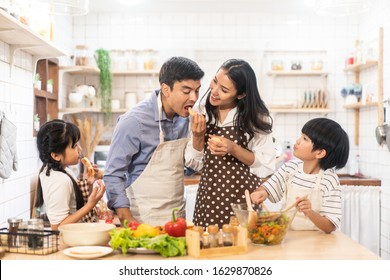 Lovely Cute Asian Family Making Food In Kitchen At Home. Portrait Of Smiling Mother, Dad And Children Standing At Cooking Counter. Mom Feeding Dad Some Fruit With Smile. Happy Family Activity Together