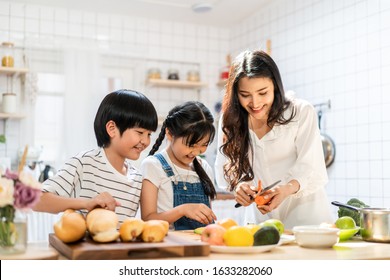 Lovely Cute Asian Family Making Food In Kitchen At Home. Portrait Of Smiling Mother And Children Standing At Cooking Counter Preparing Ingredient For Dinner Meal. Happy Family Activity Together.
