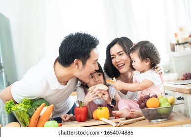 Lovely Cute Asian Family Making Food In Kitchen At Home. Portrait Of Smiling Mother, Dad And Children Standing At Cooking Counter. Kid Feeding Dad Some Bread With Smile. Happy Family Activity Together