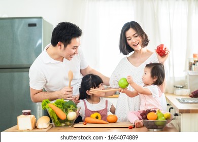 Lovely Cute Asian Family Making Food In Kitchen At Home. Portrait Of Smiling Mother, Dad And Children Standing At Cooking Counter. Kid Feeding Dad Some Fruit With Smile. Happy Family Activity Together