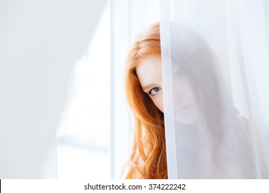 Lovely Cute Young Woman With Long Red Hair Hiding Behind White Curtains